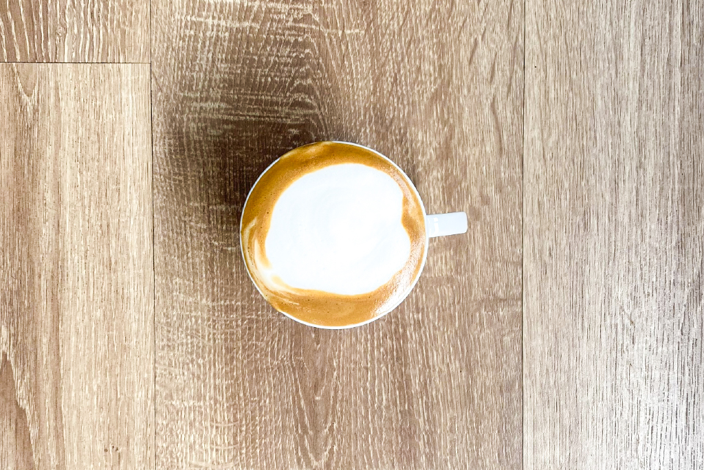 T&aacute;ch c&agrave; ph&ecirc; cappuccino nh&igrave;n từ tr&ecirc;n xuống, đặt tr&ecirc;n b&agrave;n gỗ m&agrave;u s&aacute;ng.