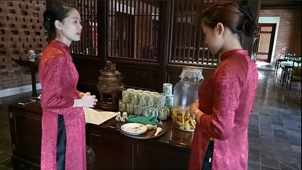 Hai người mặc áo dài đỏ đứng cạnh bàn chứa lọ thủy tinh và bộ ấm chén trong không gian truyền thống.