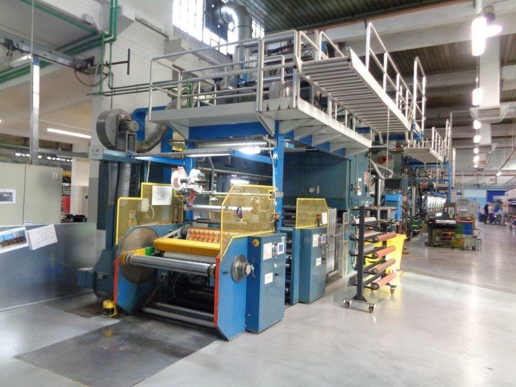 Dây chuyền máy móc công nghiệp lớn trong nhà máy, có cầu thang và hệ thống cuộn vật liệu trên sàn.