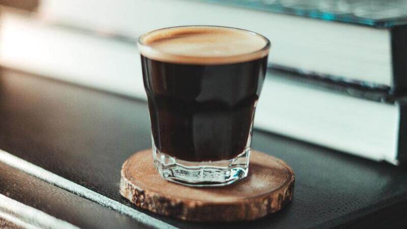 Ly cà phê espresso đặt trên đế gỗ, nền là vài quyển sách.