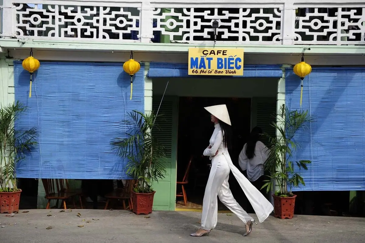 Người phụ nữ mặc áo dài trắng và nón lá đi ngang qua quán cà phê với cửa xanh và đèn lồng vàng.