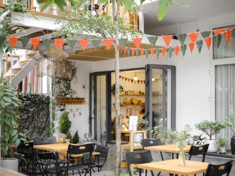 Quán cà phê ngoài trời với bàn ghế, cây xanh và dây cờ trang trí màu cam xanh, cửa kính mở ra không gian bên trong.