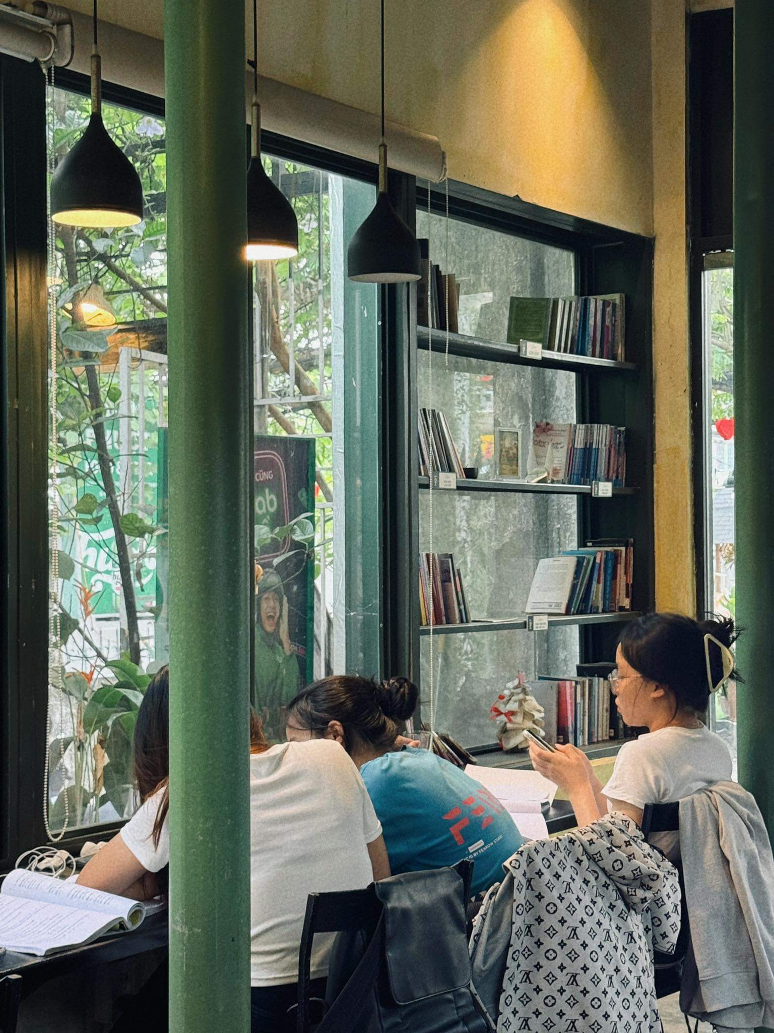 Ba người ngồi trong quán cà phê bên cửa sổ kính, đọc sách và sử dụng điện thoại, có kệ sách phía sau.