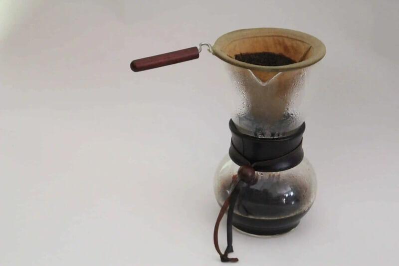 B&igrave;nh pha c&agrave; ph&ecirc; thủ c&ocirc;ng với phin vải v&agrave; tay cầm gỗ, b&ecirc;n trong c&oacute; b&atilde; c&agrave; ph&ecirc;, đặt tr&ecirc;n nền trắng.