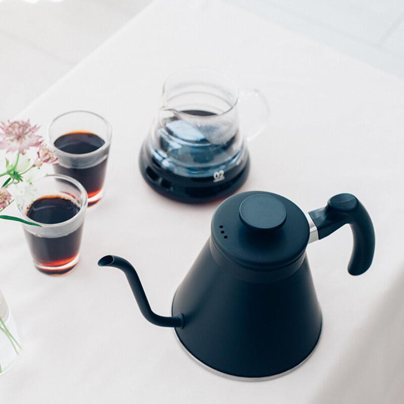 nel-drip-coffee-7 Ấm đun nước màu đen và bình chứa cà phê trên bàn, bên cạnh hai ly cà phê và một bình hoa.