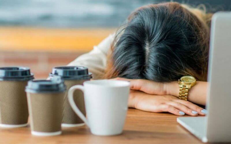 Người ngồi gục đầu trên bàn cạnh máy tính xách tay, với nhiều cốc cà phê xung quanh.