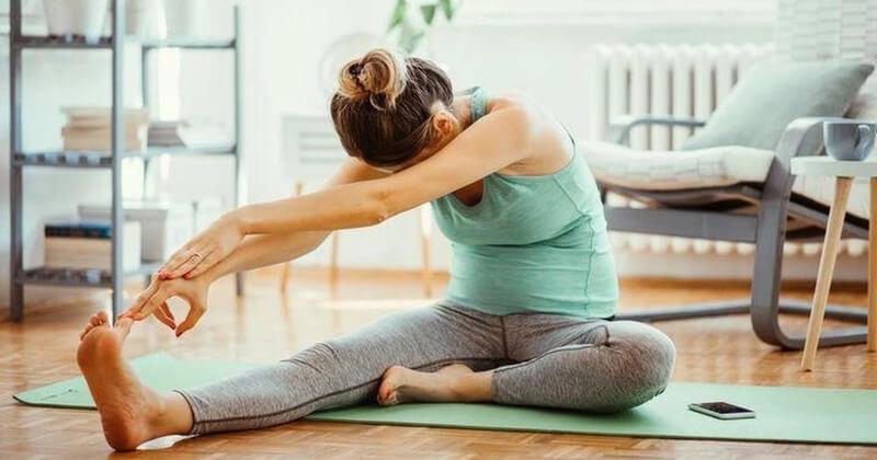 Người phụ nữ tập yoga trên thảm xanh trong phòng khách, với điện thoại bên cạnh, ánh sáng tự nhiên chiếu qua cửa sổ.