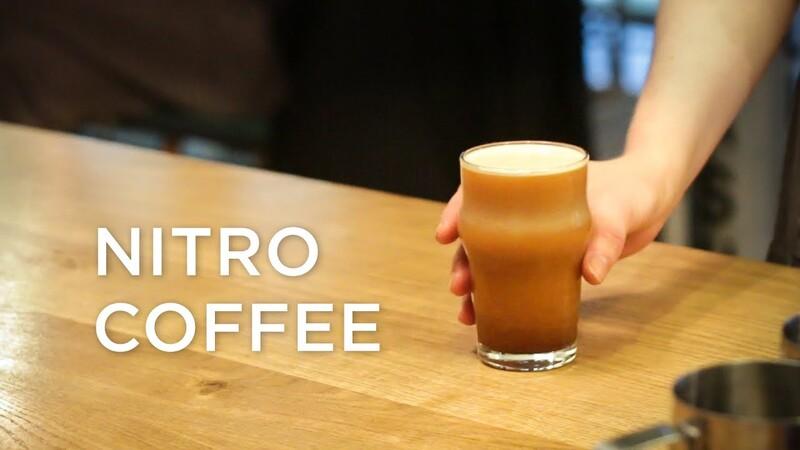 Một ly c&agrave; ph&ecirc; nitro đang được cầm tr&ecirc;n b&agrave;n gỗ, b&ecirc;n cạnh d&ograve;ng chữ "Nitro Coffee".