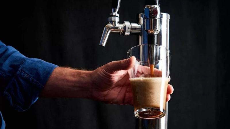 Tay người đang r&oacute;t bia từ v&ograve;i kim loại v&agrave;o ly thủy tinh, nền tối.