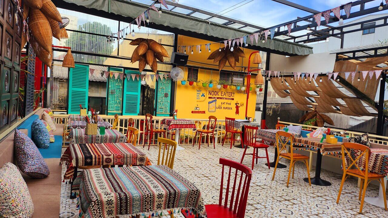 Không gian quán ăn ngoài trời với bàn ghế nhiều màu sắc, trang trí đèn và cờ, tường vàng nổi bật.