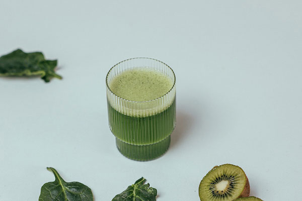 Ly nước ép xanh bên cạnh lá rau và lát kiwi trên nền trắng.