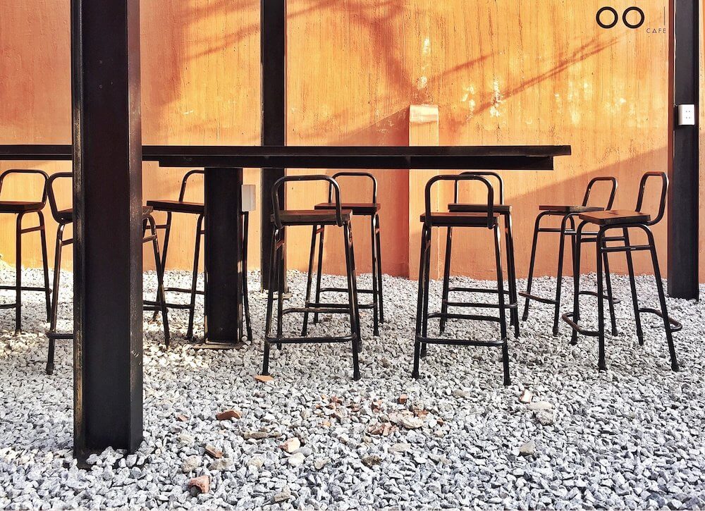 Hàng ghế cao xếp quanh bàn dài trong quán cà phê, nền tường cam và sàn rải sỏi trắng.