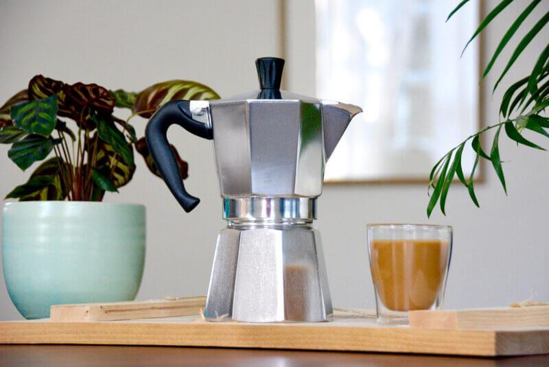 Ấm pha c&agrave; ph&ecirc; moka đặt tr&ecirc;n khay gỗ c&ugrave;ng ly c&agrave; ph&ecirc;, b&ecirc;n cạnh l&agrave; chậu c&acirc;y trong ph&ograve;ng s&aacute;ng sủa.