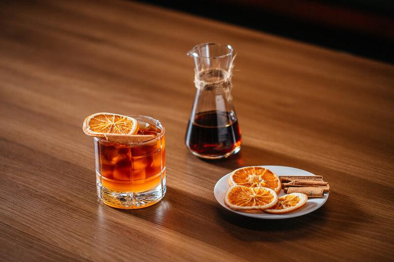 Ly cocktail với l&aacute;t cam, b&igrave;nh rượu v&agrave; đĩa cam kh&ocirc;, quế tr&ecirc;n b&agrave;n gỗ.