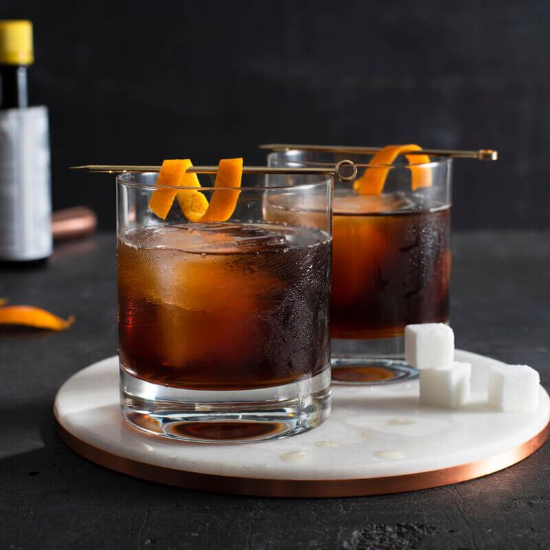 Hai ly cocktail với đ&aacute;, trang tr&iacute; vỏ cam v&agrave; xi&ecirc;n kim loại, đặt tr&ecirc;n đế tr&ograve;n, cạnh v&agrave;i vi&ecirc;n đường.