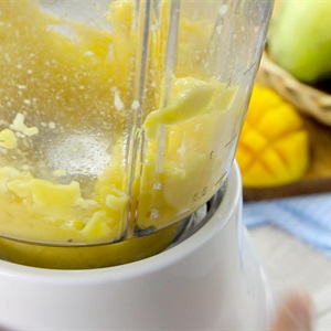 M&aacute;y xay sinh tố đang xay hỗn hợp m&agrave;u v&agrave;ng, c&oacute; thể l&agrave; xo&agrave;i hoặc tr&aacute;i c&acirc;y kh&aacute;c, tr&ecirc;n b&agrave;n c&oacute; khăn kẻ &ocirc;.