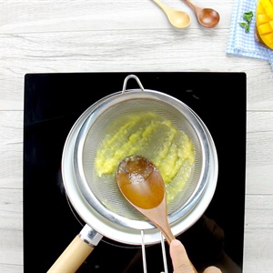 Người d&ugrave;ng lọc hỗn hợp m&agrave;u v&agrave;ng qua r&acirc;y tr&ecirc;n bếp, nền gỗ trắng, c&oacute; th&igrave;a gỗ v&agrave; khăn kẻ caro.