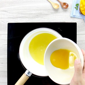 Người đang đổ chất lỏng m&agrave;u v&agrave;ng từ b&aacute;t v&agrave;o chảo tr&ecirc;n bếp, b&ecirc;n cạnh l&agrave; một số dụng cụ nh&agrave; bếp.