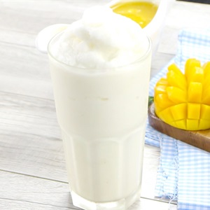 Ly sinh tố trắng mịn tr&ecirc;n b&agrave;n gỗ, b&ecirc;n cạnh l&agrave; miếng xo&agrave;i v&agrave;ng tươi v&agrave; khăn caro xanh trắng.