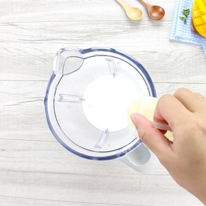 Một tay đang đổ chất lỏng m&agrave;u trắng v&agrave;o cối xay sinh tố tr&ecirc;n b&agrave;n gỗ, b&ecirc;n cạnh c&oacute; tr&aacute;i xo&agrave;i v&agrave; th&igrave;a gỗ.