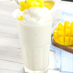 Ly sinh tố trắng với topping xo&agrave;i cắt nhỏ, b&ecirc;n cạnh l&agrave; đĩa xo&agrave;i ch&iacute;n được cắt l&aacute;t trang tr&iacute; tr&ecirc;n khăn sọc xanh.