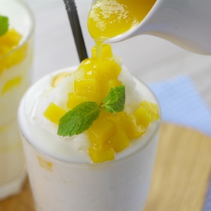 Ly sinh tố trắng c&oacute; đ&aacute; v&agrave; xo&agrave;i, trang tr&iacute; l&aacute; bạc h&agrave;, đang được r&oacute;t th&ecirc;m nước xo&agrave;i từ một b&igrave;nh nhỏ.