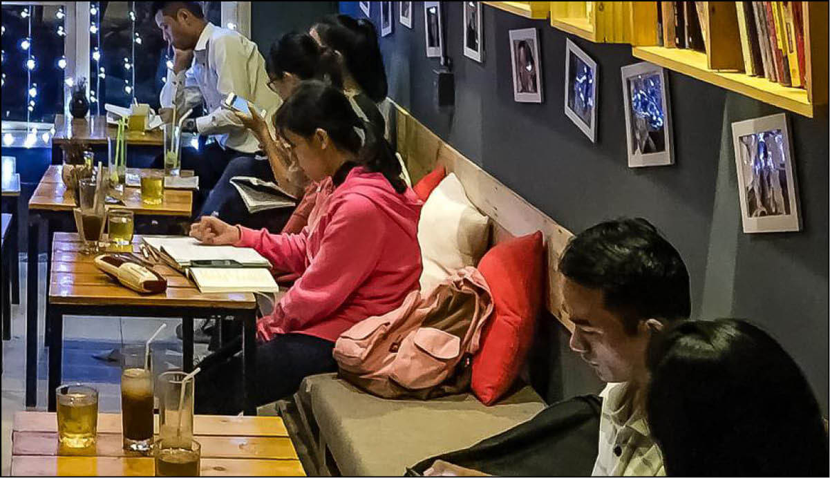 Quán cà phê với nhiều người ngồi đọc sách và sử dụng điện thoại, trên bàn có đồ uống và sách.