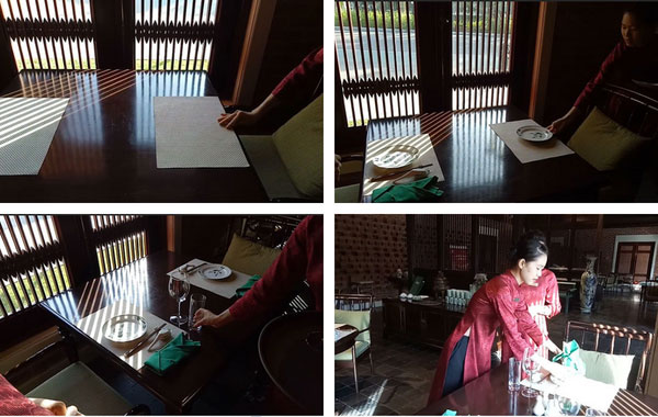 Người đang sắp xếp bàn ăn trong nhà hàng, có ánh sáng chiếu qua cửa sổ, với đĩa và ly đặt trên bàn.