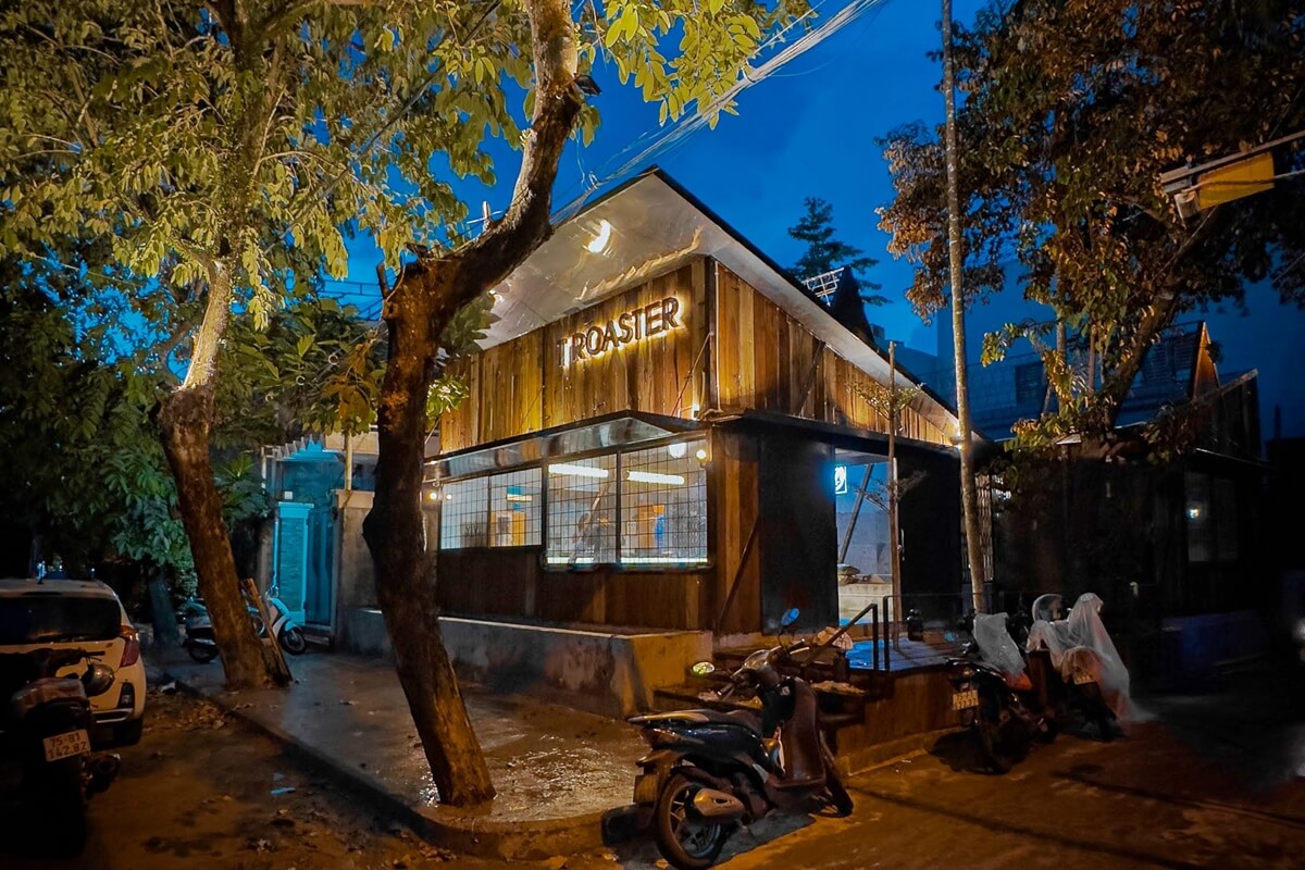 Quán cà phê với ánh sáng ấm áp vào buổi tối, xung quanh có cây cối và xe máy đậu bên lề đường.