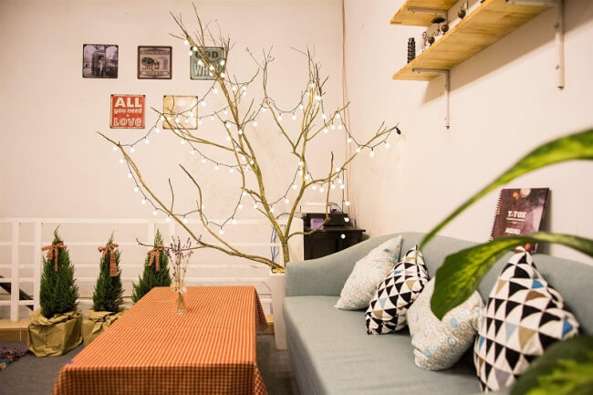 Phòng khách với ghế sofa xanh, bàn trải khăn caro, cây trang trí đèn nhấp nháy và kệ gỗ treo tường.