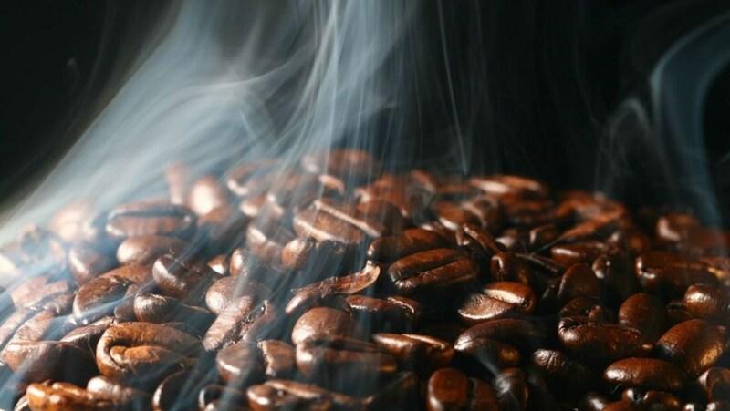 Hạt cà phê rang đang bốc khói, tạo ra làn khói mỏng bay lên trong không khí nền tối.