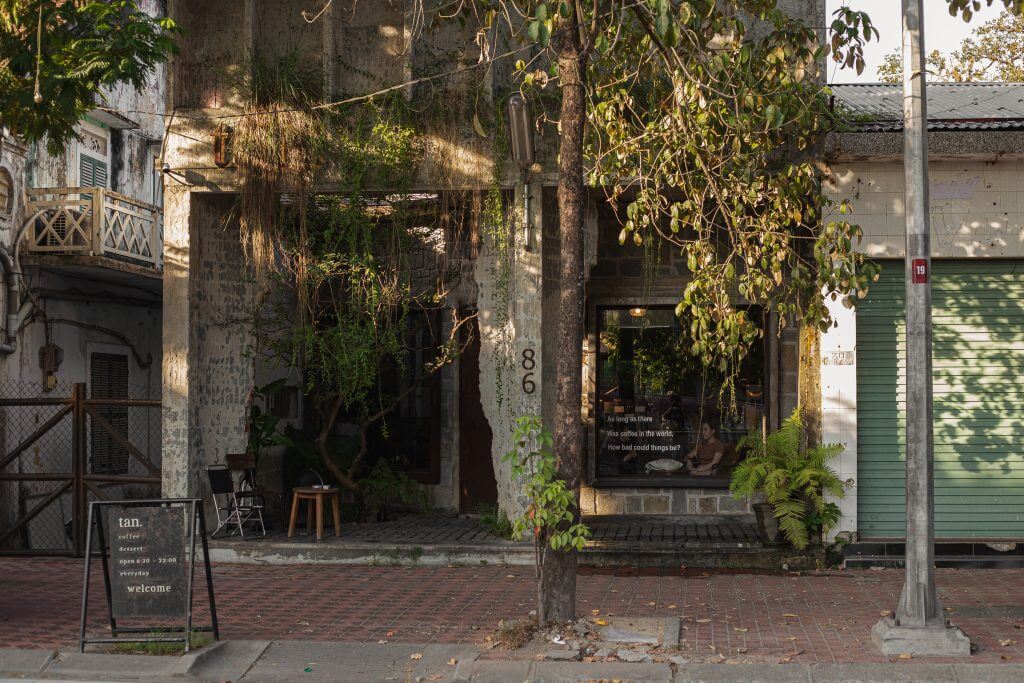 Mặt tiền quán cà phê với cây xanh bao phủ, biển hiệu và cửa cuốn màu xanh lá cây bên cạnh.