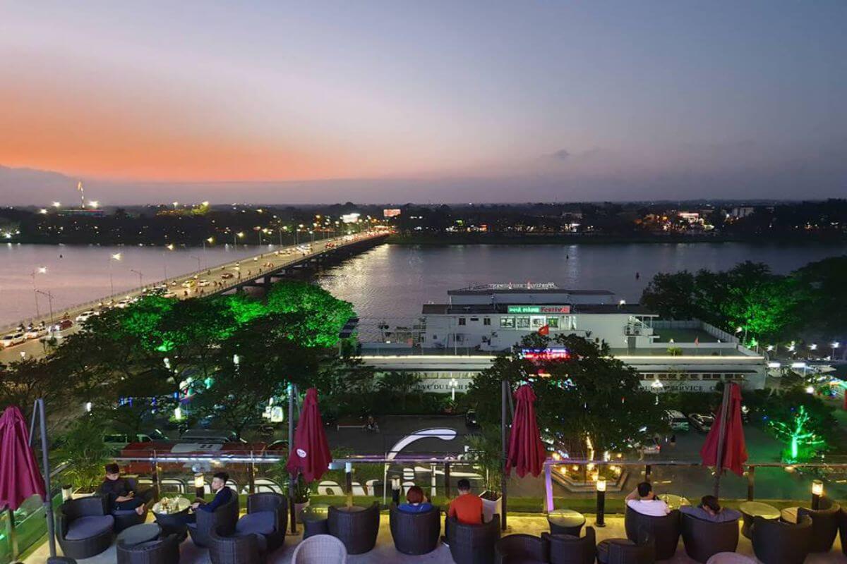 Khung cảnh hoàng hôn bên sông, với cây cầu và một quán cà phê có khách ngồi thư giãn ngoài trời.