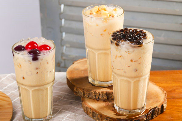 Ba ly trà sữa đá, mỗi ly có topping khác nhau gồm trân châu đen, trái cây và thạch, đặt trên bàn gỗ.