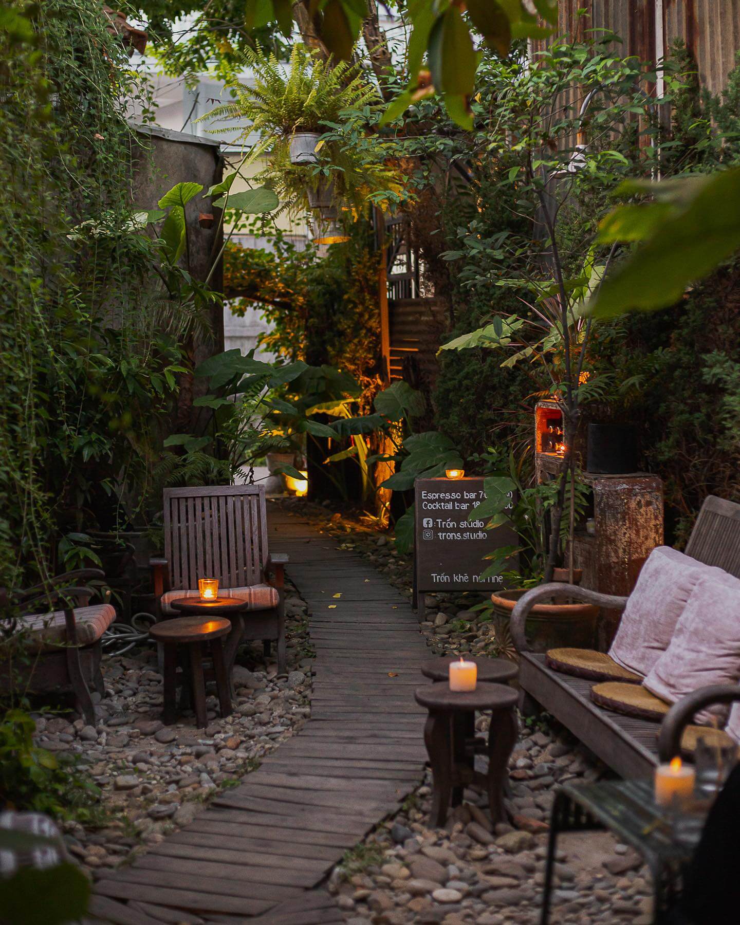 Lối đi lát gỗ dẫn qua khu vườn xanh mát, có bàn ghế gỗ và ánh nến lung linh tạo không gian thư giãn.