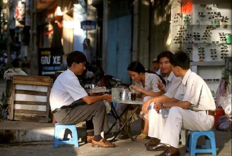 van-hoa-ca-phe-viet-nam-9 Bốn người ngồi uống cà phê ngoài trời trên ghế nhựa xanh, trò chuyện cạnh kệ trưng bày kính mát.