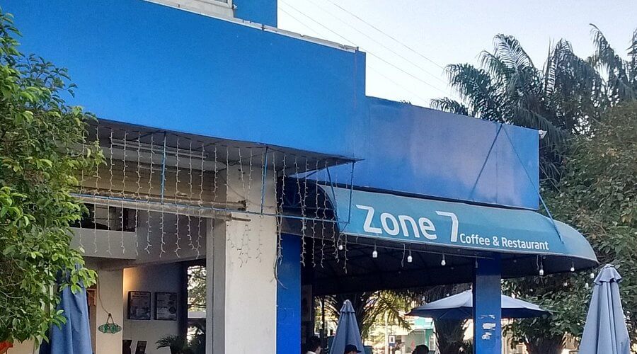 Quán cà phê và nhà hàng với bảng hiệu "Zone 7", bên ngoài có cây xanh và ô che nắng.