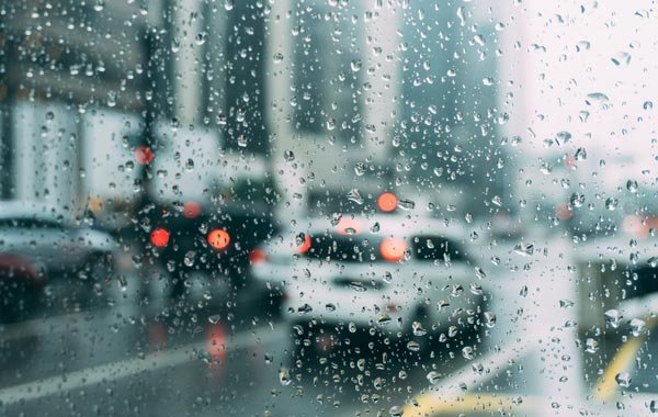 Kính xe có giọt nước mưa, phía sau là cảnh đường phố với xe cộ mờ nhạt và đèn xe sáng trong thời tiết mưa.