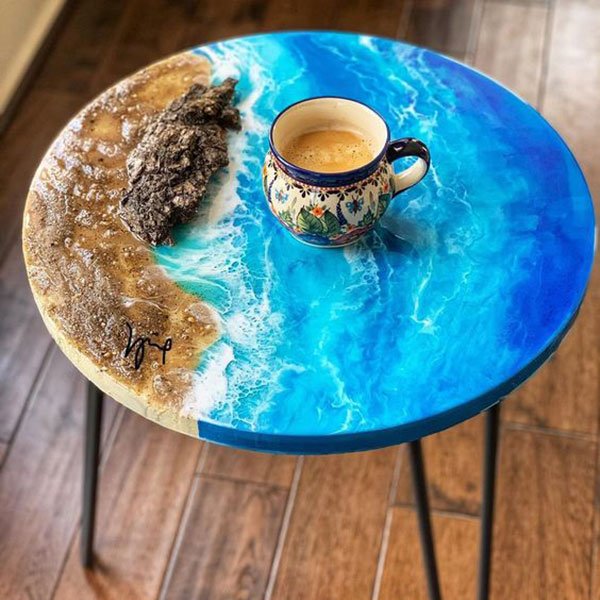 Bàn tròn với mặt bàn trang trí hình bờ biển và sóng biển, có một tách cà phê đặt trên bề mặt.