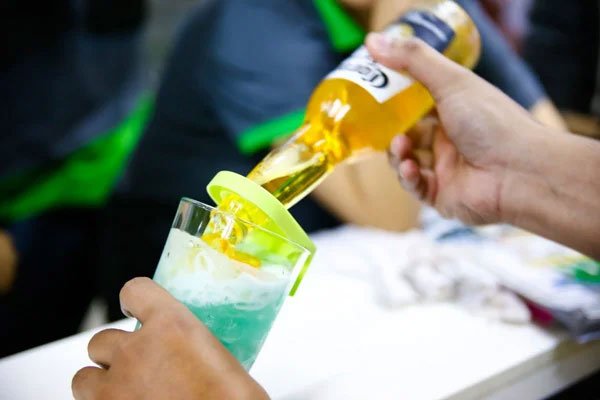 Người đang rót bia từ chai vào ly cocktail màu xanh, có nắp nhựa màu vàng trên miệng ly.