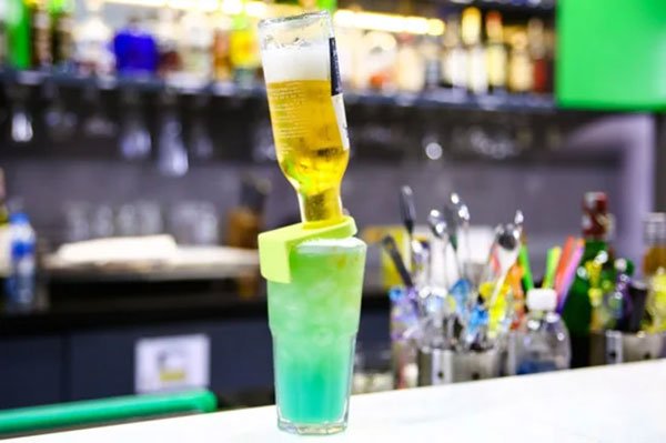 Ly cocktail màu xanh với chai bia úp ngược trên quầy bar, nền có nhiều chai lọ và dụng cụ pha chế.