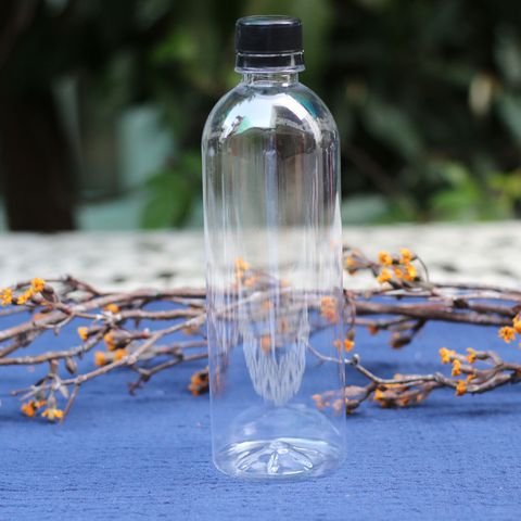 Chai nhựa trong suốt c&oacute; nắp đen đặt tr&ecirc;n nền vải xanh, xung quanh l&agrave; c&agrave;nh c&acirc;y kh&ocirc; với hoa nhỏ m&agrave;u cam.