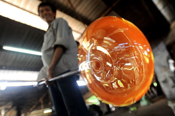 Người thổi thuỷ tinh đang tạo hình một khối thuỷ tinh nóng chảy màu cam trong xưởng sản xuất.