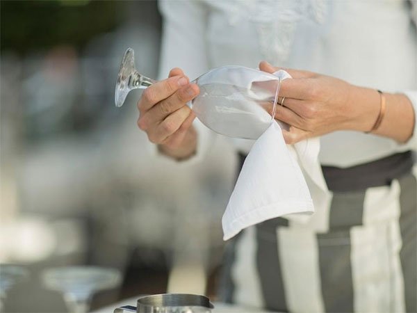 dung-khan-mem-lau-do-thuy-tinh Người đang lau ly rượu bằng khăn trắng, mặc áo sáng màu, trong không gian mờ nhạt.