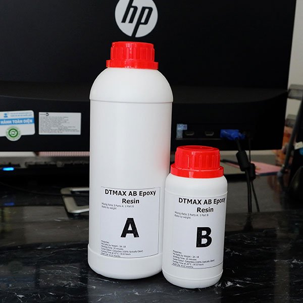 Hai chai nhựa với nắp đỏ, nhãn ghi "DTMAX AB Epoxy Resin", đặt trước màn hình máy tính.