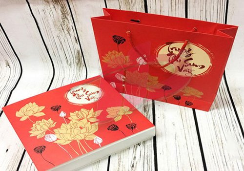 Hộp qu&agrave; v&agrave; t&uacute;i m&agrave;u đỏ với họa tiết hoa sen v&agrave; chữ viết tay tr&ecirc;n nền gỗ trắng.