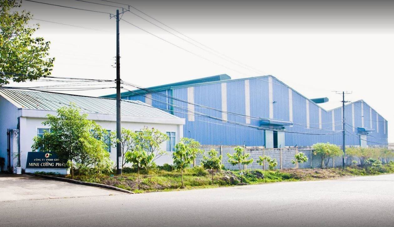 Khu nh&agrave; xưởng với m&aacute;i t&ocirc;n xanh, b&ecirc;n ngo&agrave;i c&oacute; c&acirc;y xanh v&agrave; đường đi ph&iacute;a trước, trời nắng s&aacute;ng.