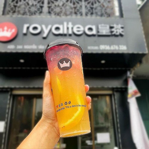 Ly tr&agrave; sữa nhiều m&agrave;u sắc được cầm tr&ecirc;n tay, nền l&agrave; bảng hiệu của một qu&aacute;n tr&agrave;.