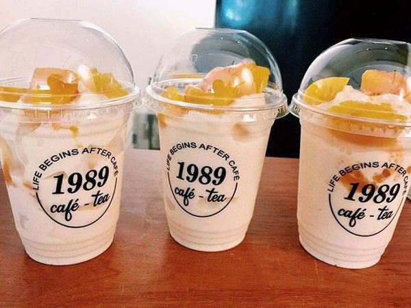 Ba ly đồ uống trắng có nắp nhựa trong suốt, trên bàn gỗ, trang trí với trái cây và có dòng chữ trên ly.