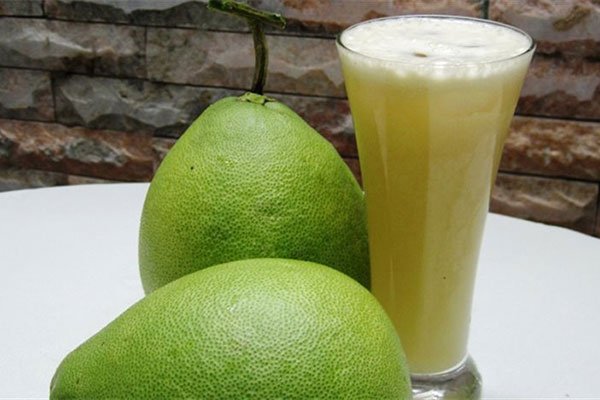 Hai quả bưởi xanh b&ecirc;n cạnh ly nước &eacute;p bưởi m&agrave;u v&agrave;ng nhạt tr&ecirc;n b&agrave;n trắng, nền l&agrave; bức tường gạch.